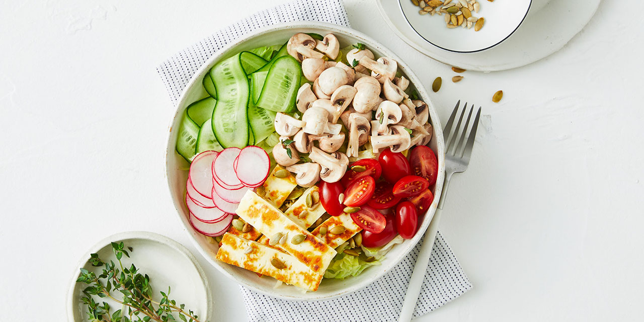 A poke bowl on a white table with mushrooms, cucumber, tomatoes, radishes and haloumi